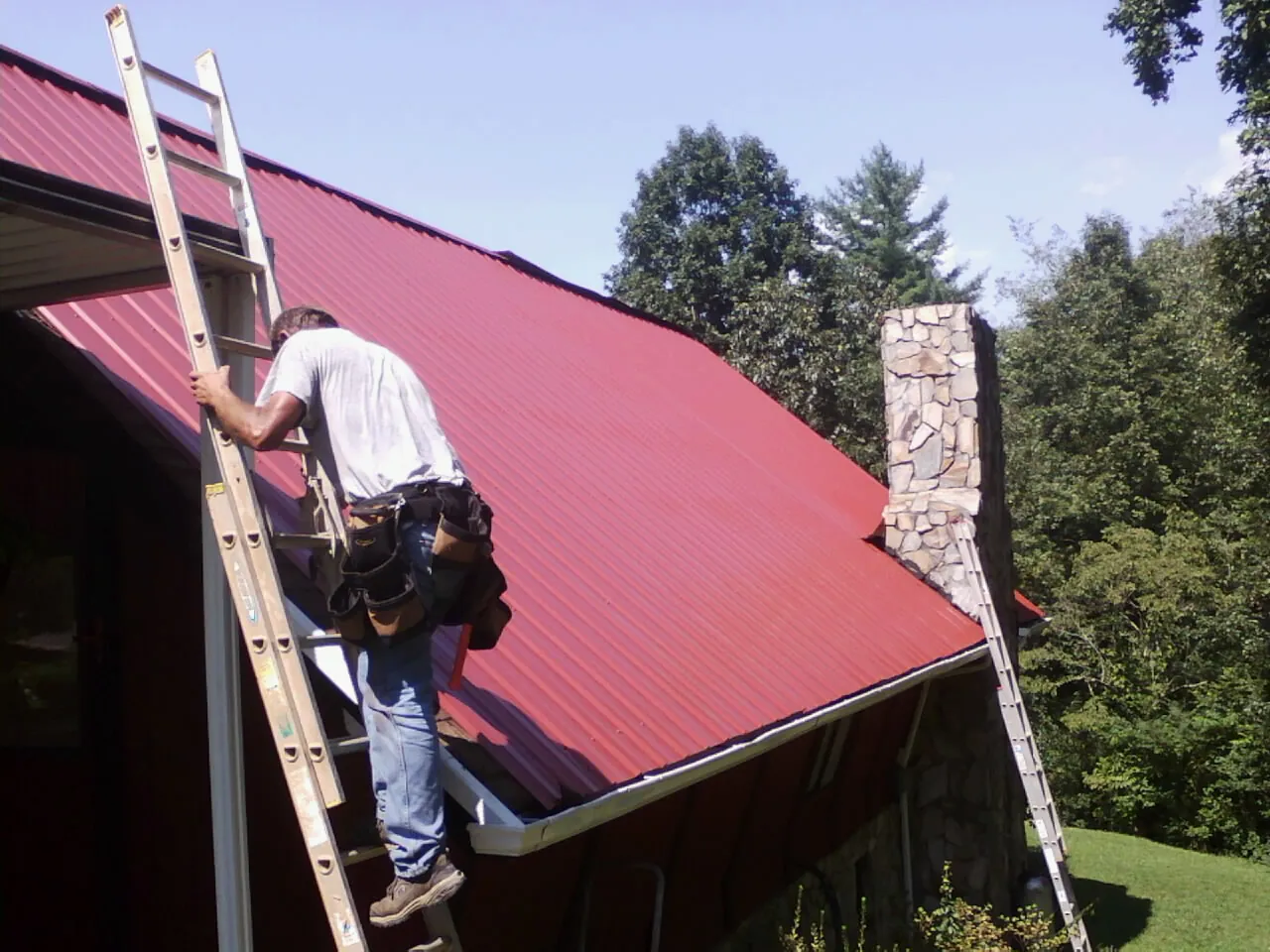 Roofer on a ladder beside a red metal roof and stone chimney — Soffit & Fascia Repair by Roofing Brothers in Allison Park